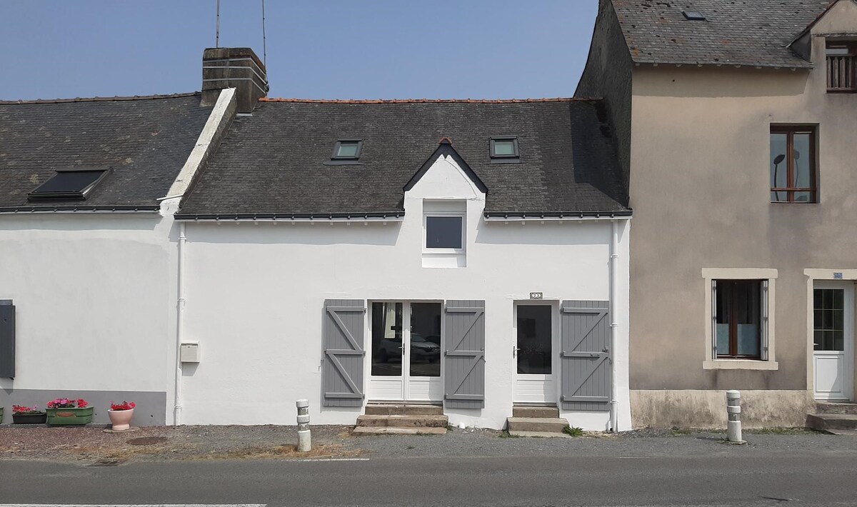 The exterior of a white, renovated house is presented, featuring grey shutters and a central window above the front door. Steps lead up to the entrance, with a simple pathway bordered by gravel. Neighbouring buildings are visible, creating a charming street view.