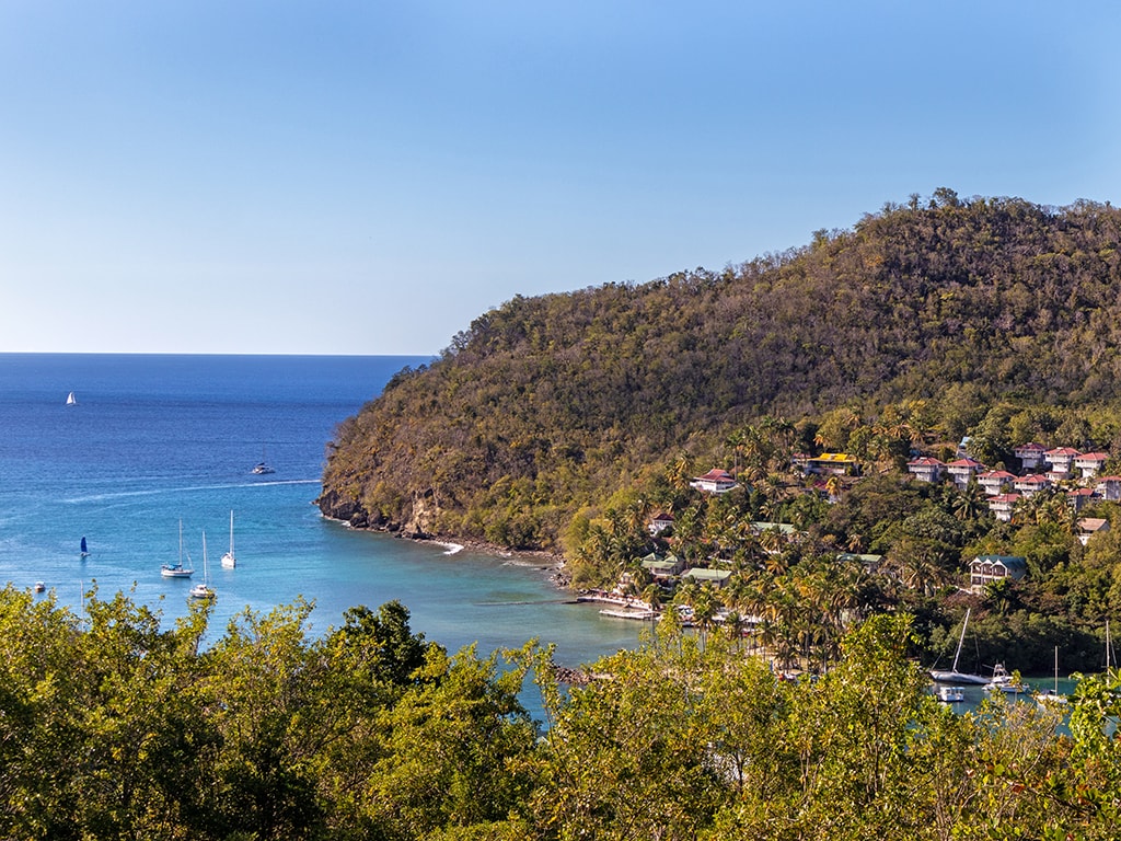 A scenic view of Marigot Bay features lush green hillsides meeting the clear blue water. Sailboats are anchored in the tranquil bay, while distant coastal buildings are nestled among the trees. The sky is bright and clear, enhancing the natural beauty of the area.
