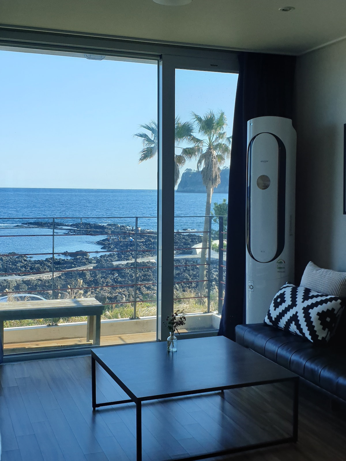 A living area is viewed through large glass doors, offering a direct sight of the ocean and nearby palm trees. A black sofa is positioned across from a simple coffee table, while a decorative pillow adds a touch of comfort. The coastal scenery is framed beautifully, enhancing the room's natural light.