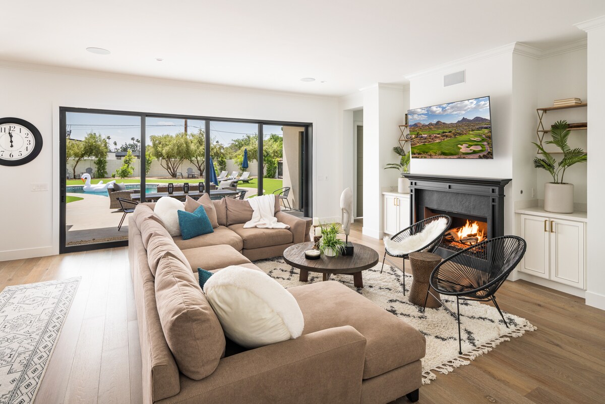A spacious living area features a large sectional sofa arranged around a central coffee table, with a cozy gas fireplace beneath a mounted TV. Floor-to-ceiling glass doors provide views of the outdoor pool area, while natural light fills the room.