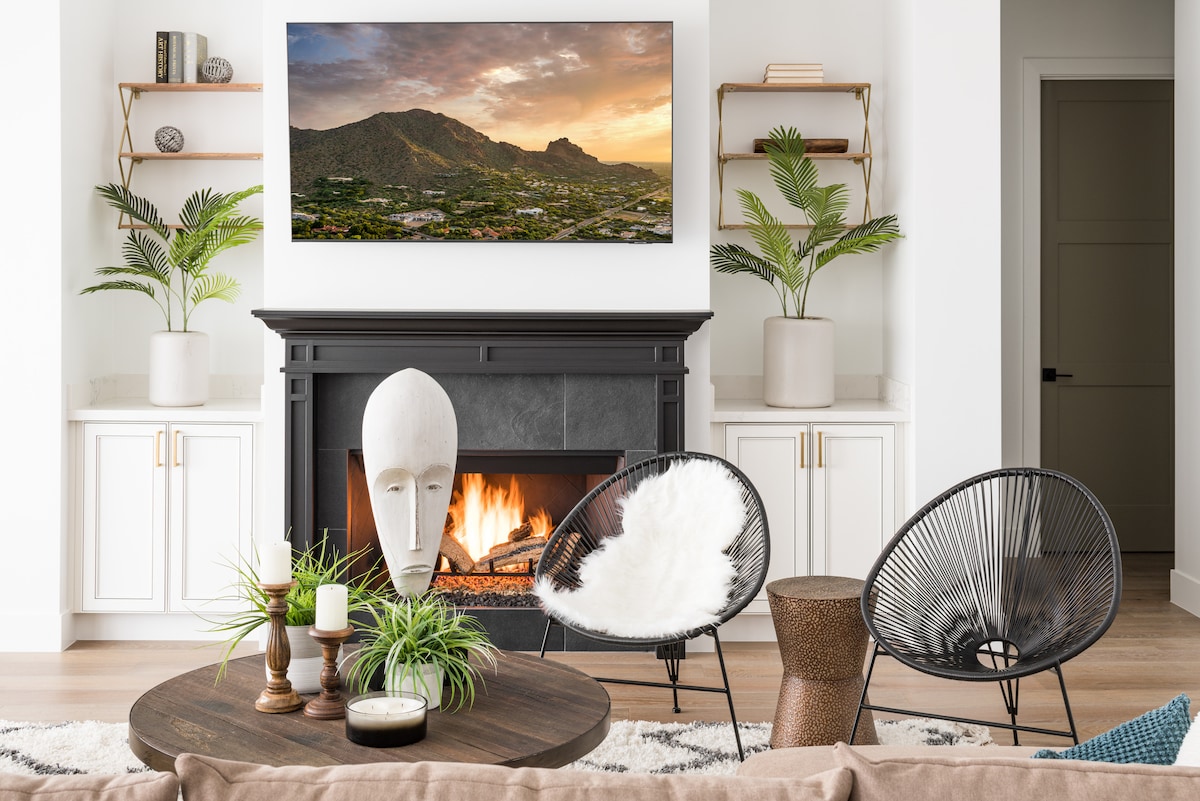 A modern living space features a black fireplace with a warm fire, flanked by two stylish chairs. A round coffee table, adorned with plants and candles, complements the room’s neutral tones. A large artwork depicting a mountainous landscape hangs above the fireplace.