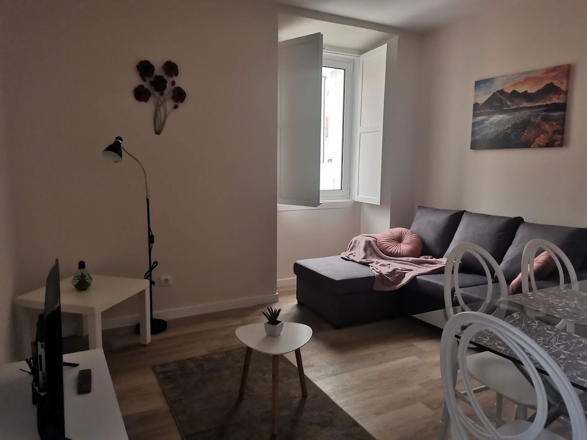 A well-lit living area features a comfortable sectional sofa adorned with decorative cushions. A small round coffee table sits on the wooden floor, while a dining table with clear chairs is visible in the background. A painting of mountains adorns one wall, and sunlight streams through open windows.