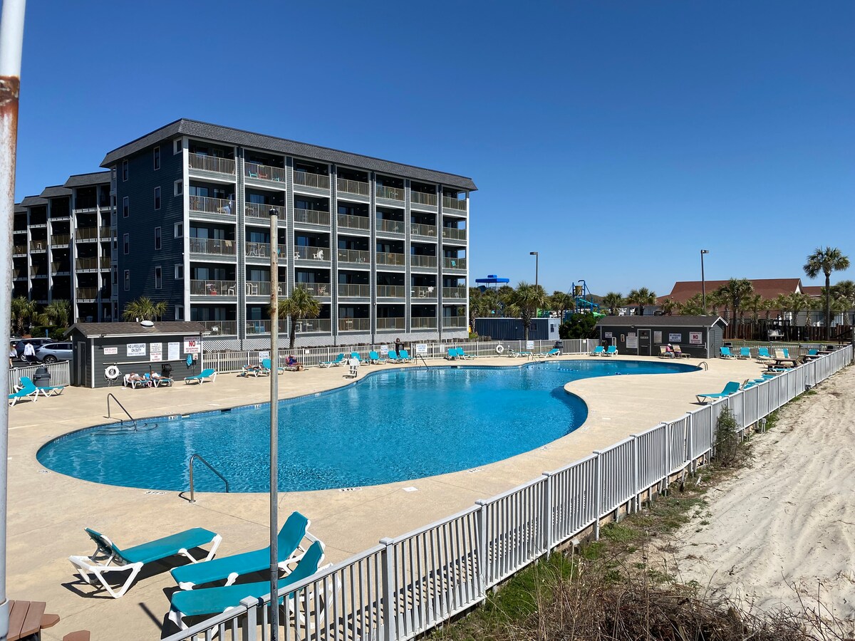 An expansive outdoor pool is framed by lounge chairs, providing ample space for relaxation. The surrounding area features palm trees and a sandy path leading to additional resort amenities, with the multi-story building visible in the background under a clear blue sky.