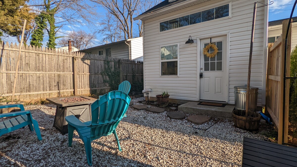 A private outdoor space is displayed, featuring gravel flooring and a fire pit surrounded by Adirondack chairs. The guesthouse exterior is visible, with large windows allowing for natural light. A simple wreath adorns the front door, enhancing the cozy atmosphere of the setting.