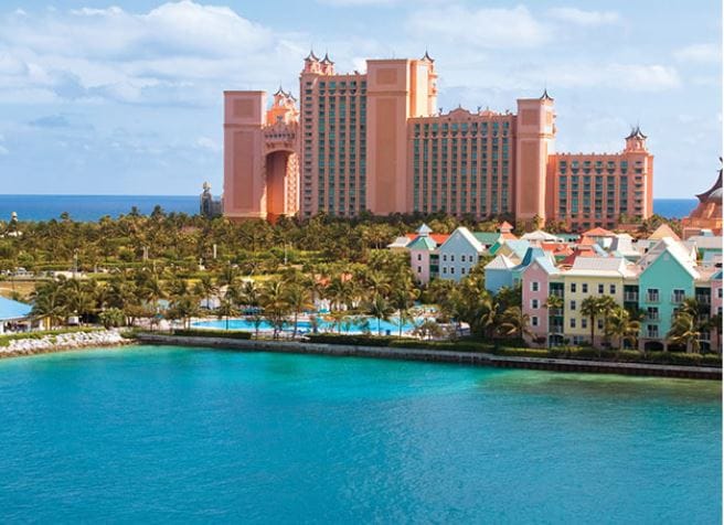 The image captures a panoramic view of the Atlantis resort's iconic pink-colored towers overlooking a vibrant turquoise sea. Lush palm trees line the waterfront, and colorful buildings dot the shoreline, complemented by recreational pools that invite relaxation.