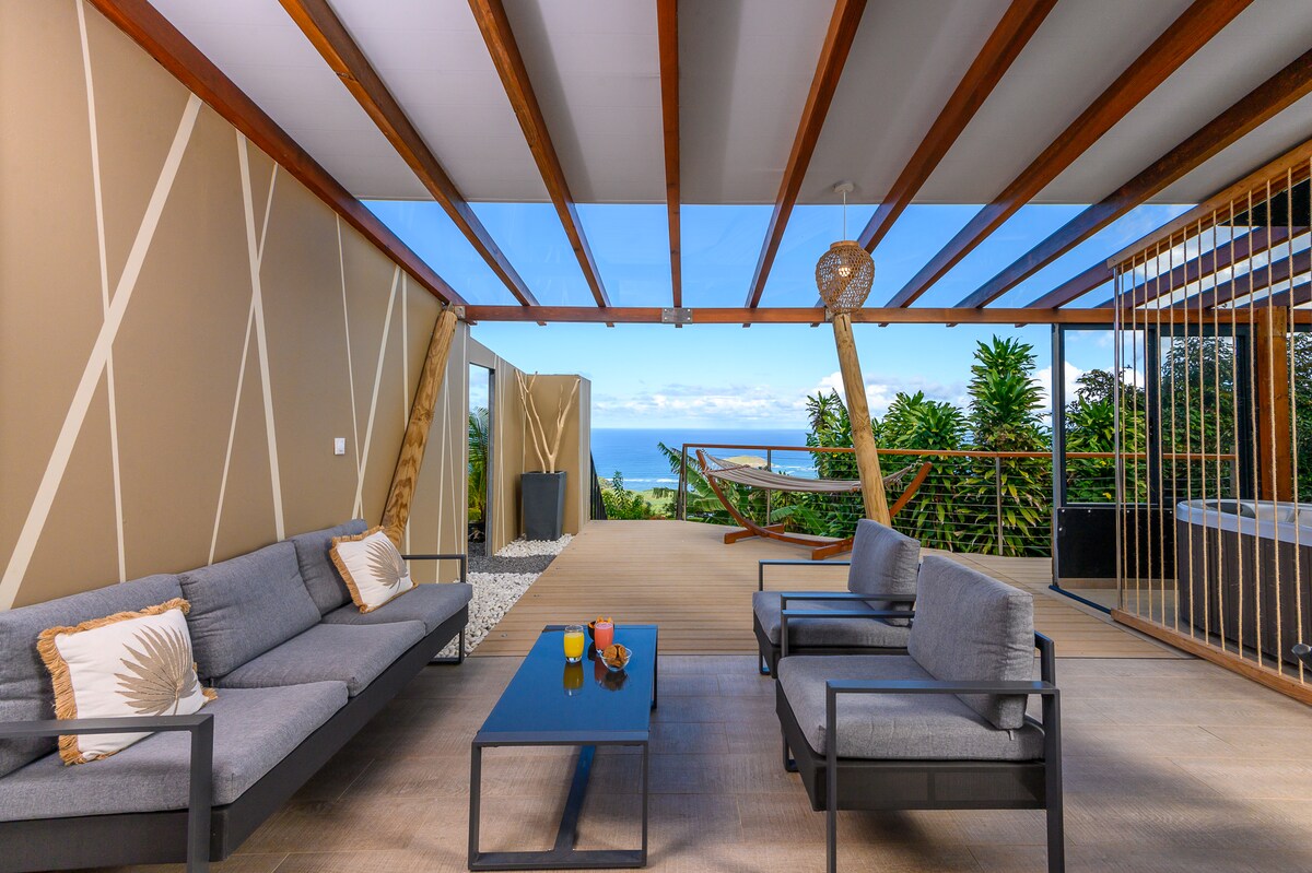An outdoor terrace features a comfortable seating area with gray sofas and a black coffee table. A hammock is positioned in the background, and a private jacuzzi is visible to the side. Scenic views of the sea and greenery are framed by wooden beams overhead.