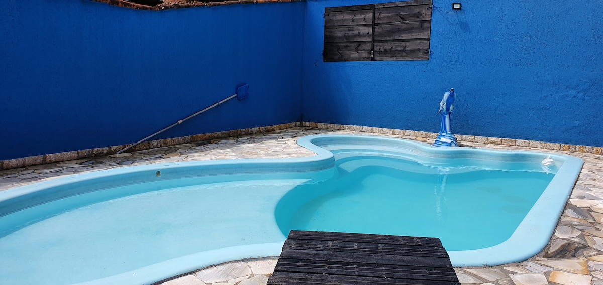 A private swimming pool features a unique shape and is surrounded by smooth stone tiles. A blue wall provides a vibrant backdrop, while a pool cleaning device rests at the edge. The water reflects the sky, contributing to a serene environment.