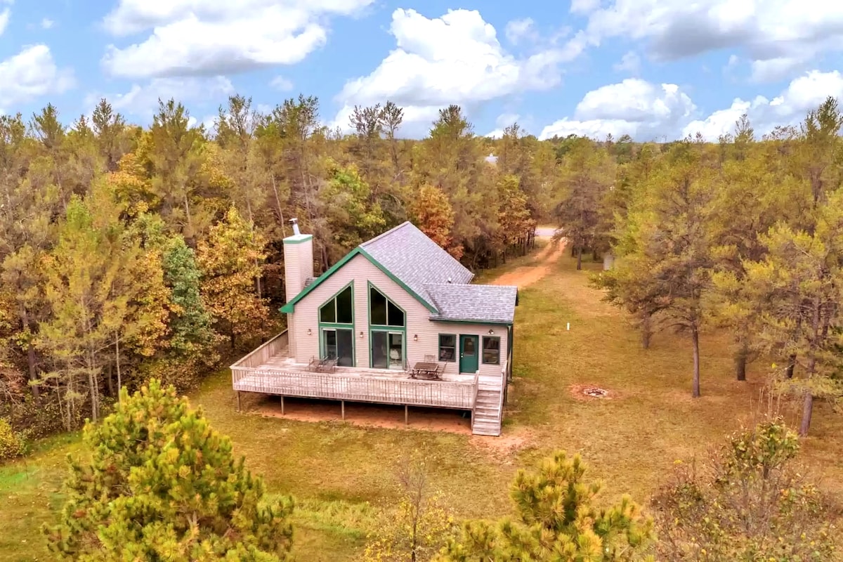 The chalet is set in a wooded area, showcasing a large deck in front. The pentagonal windows invite natural light, complemented by a stone chimney. The surrounding landscape features a mix of trees, and a clearly defined path leads toward the property.
