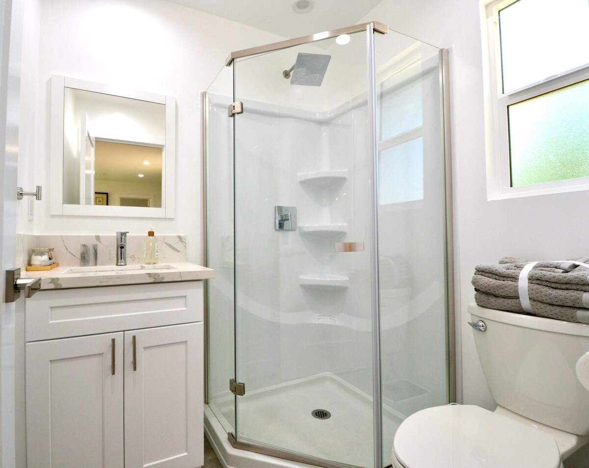A modern bathroom features a frameless glass shower with built-in shelves. A single vanity with a mirror is positioned beside the shower, and neatly rolled towels are arranged on the countertop. Natural light filters through frosted windows, enhancing the clean and bright atmosphere.