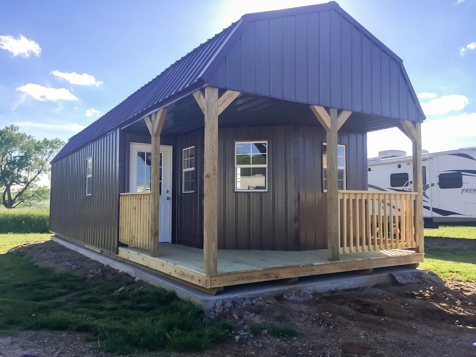 The exterior of a cottage is shown, featuring a sturdy metal roof and wooden porch with a railing. Large windows allow natural light inside, and a grassy area surrounds the structure. Nearby, additional structures are visible in the background.