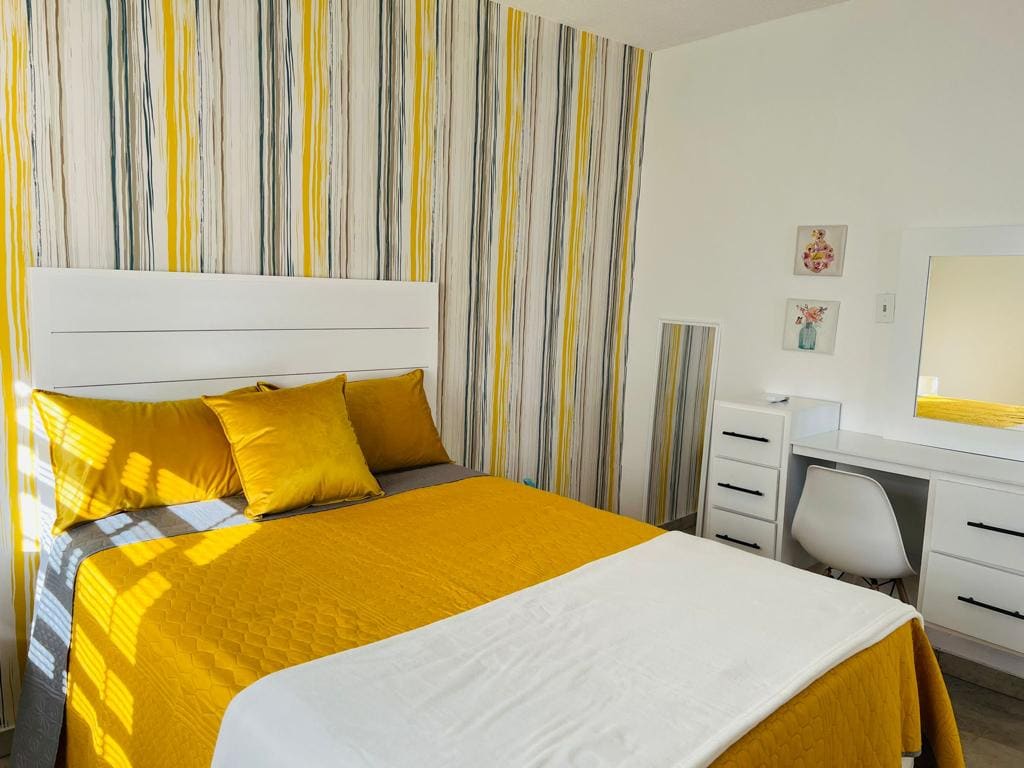 A brightly colored bedroom features a spacious bed covered with a vibrant yellow quilt and decorative pillows. A striped wall adds an artistic touch. A sleek desk and chair are positioned nearby, illuminated by natural light from a window.