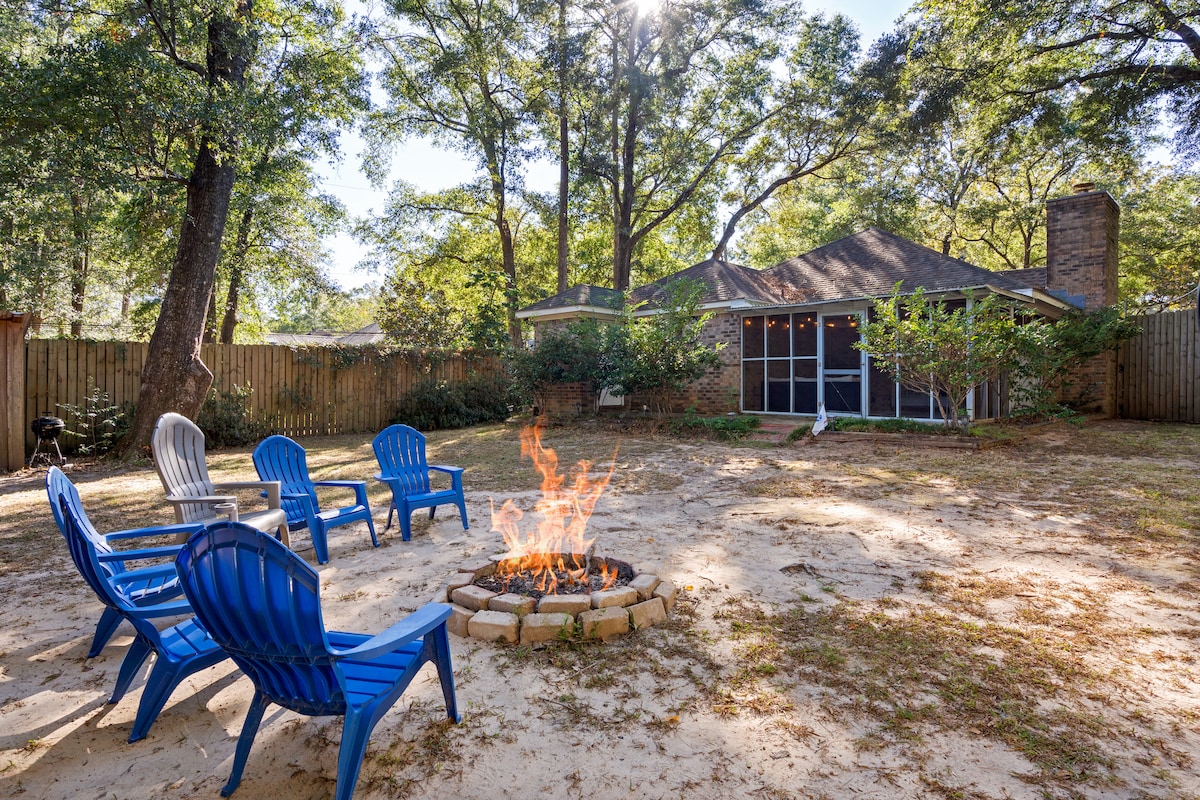 An outdoor fire pit area is surrounded by five blue Adirondack chairs positioned in a circular layout. A selection of trees and shrubbery provides a natural backdrop, while sunlight filters through the leaves, illuminating the sandy ground.