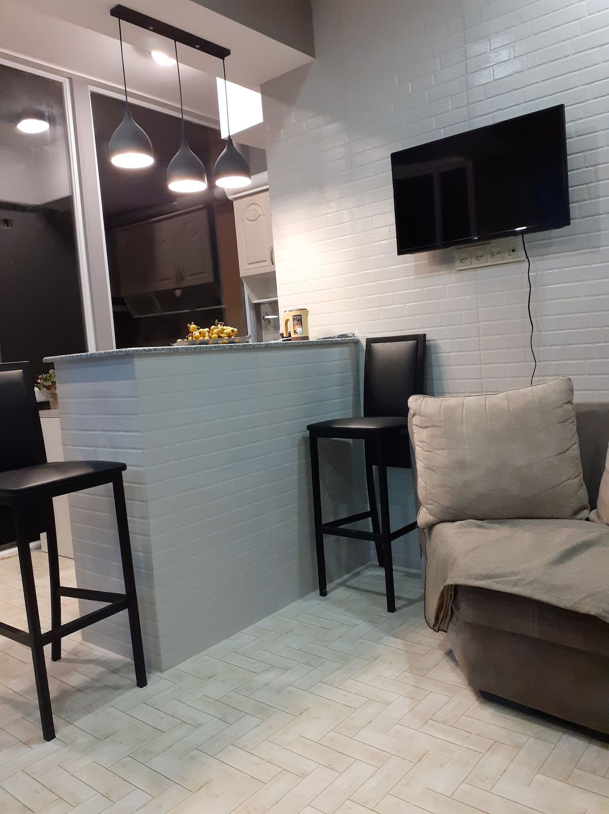 A modern kitchen area is highlighted by a white brick backsplash and sleek bar counter with high black stools. A light-colored sofa is positioned nearby, with a throw blanket draped over the arm. A flat-screen TV is mounted on the wall, adding convenience for guests.