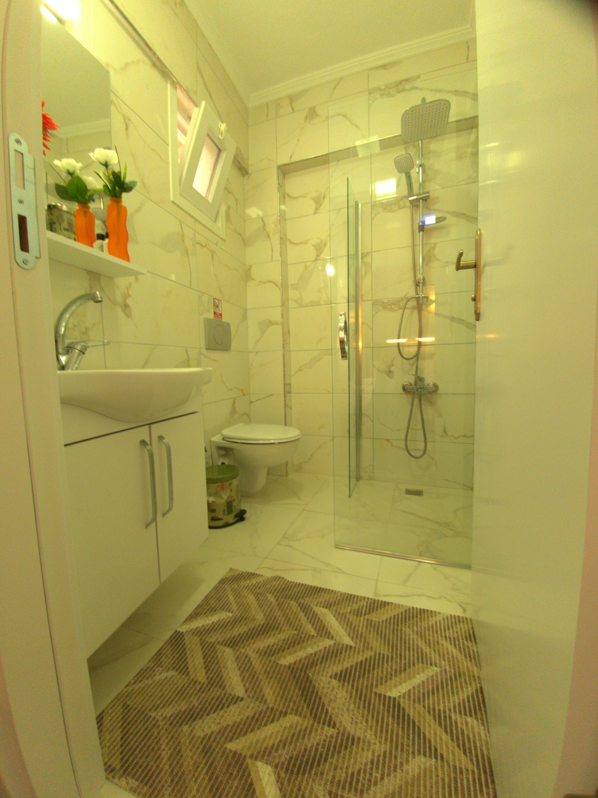 A modern bathroom is characterized by marble-style tiles and a glass-enclosed shower. Features include a contemporary sink with storage below, a wall-mounted mirror, and a sleek toilet. A patterned area rug adds texture to the space, while natural light enters through a small window.