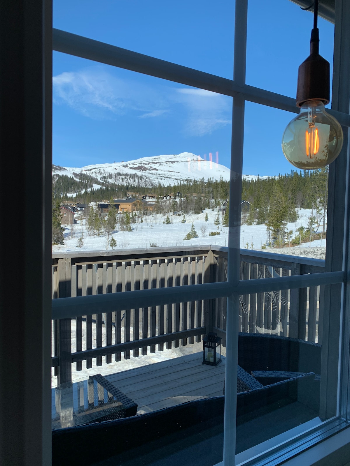 A picturesque view through a large window showcases a snow-covered landscape, featuring distant mountains and forested areas. A balcony can be seen below, which is furnished for outdoor relaxation.