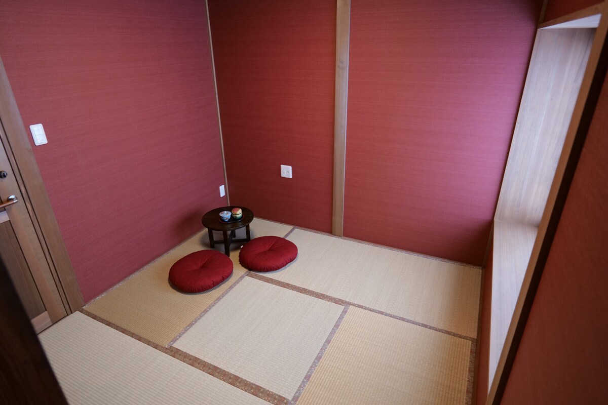 A small room with deep red-brown walls and tatami mat flooring is depicted. A low table is centered, accompanied by two round cushions. Natural light is softly filtered through a nearby window, creating a quiet and serene ambiance.