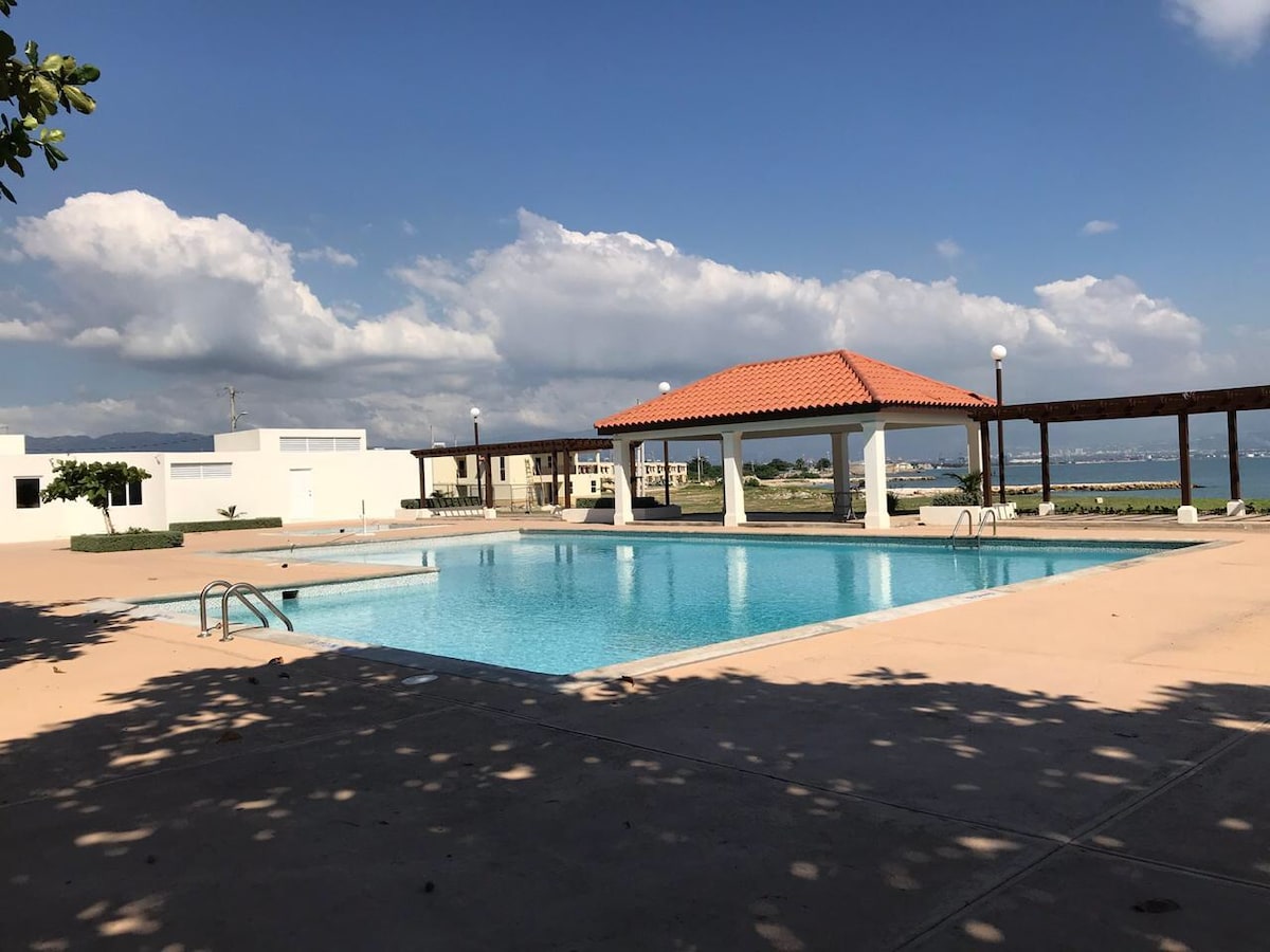 A clear blue swimming pool is surrounded by a spacious patio with shaded seating areas. The structure features a tiled gazebo with a red roof, and the serene landscape includes distant views of water and sky, contributing to a peaceful outdoor environment.
