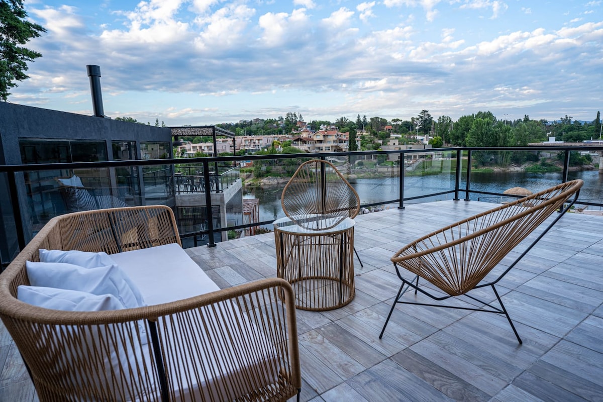 An outdoor terrace is furnished with stylish seating that includes a white sofa and two woven chairs. The surrounding area offers views of the lake and greenery, with a backdrop of a clear sky and distant residential buildings.