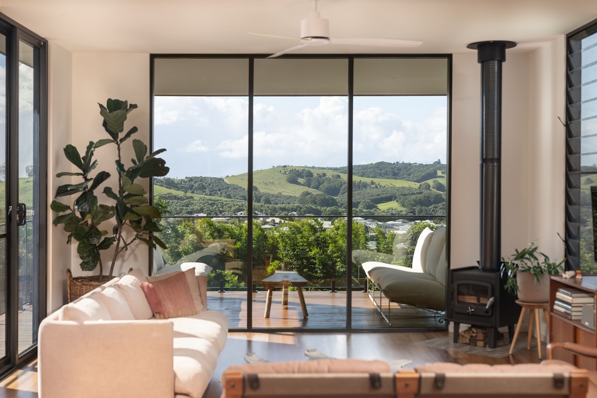 A spacious living area is presented with floor-to-ceiling glass doors that provide a view of gently rolling hills. A light-colored sofa and a cozy chair are positioned for relaxation, complemented by a small coffee table. Natural greenery enhances the inviting atmosphere.
