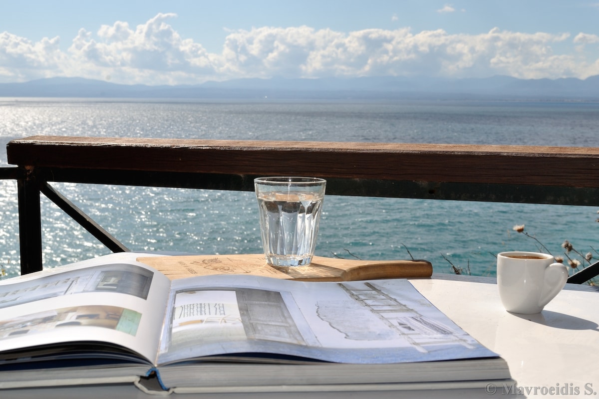 A table made of wood features an open book and a small cup of coffee beside a glass of water. The sparkling sea can be seen in the background, with clouds gently dotting the sky, creating a serene atmosphere.