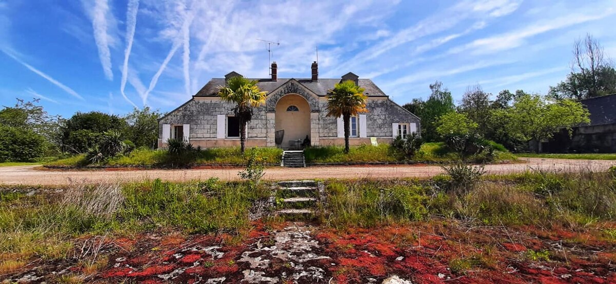 The exterior of a spacious home is presented, showcasing a symmetrical façade with an arched entryway. Lush greenery surrounds the property, complemented by colorful ground cover. The sky above is bright with wispy clouds, contributing to a serene atmosphere.