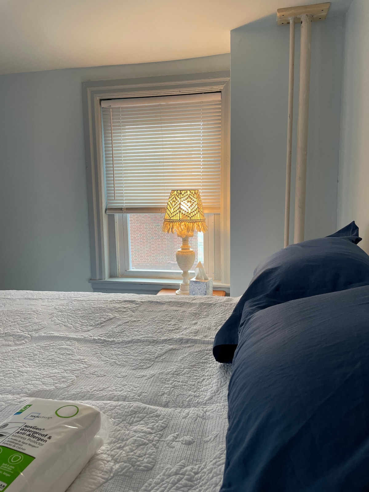 A cozy bedroom is depicted, featuring a spacious bed topped with dark blue pillows. A bedside lamp with a decorative shade provides soft illumination beside a window adorned with blinds, allowing natural light to filter through.