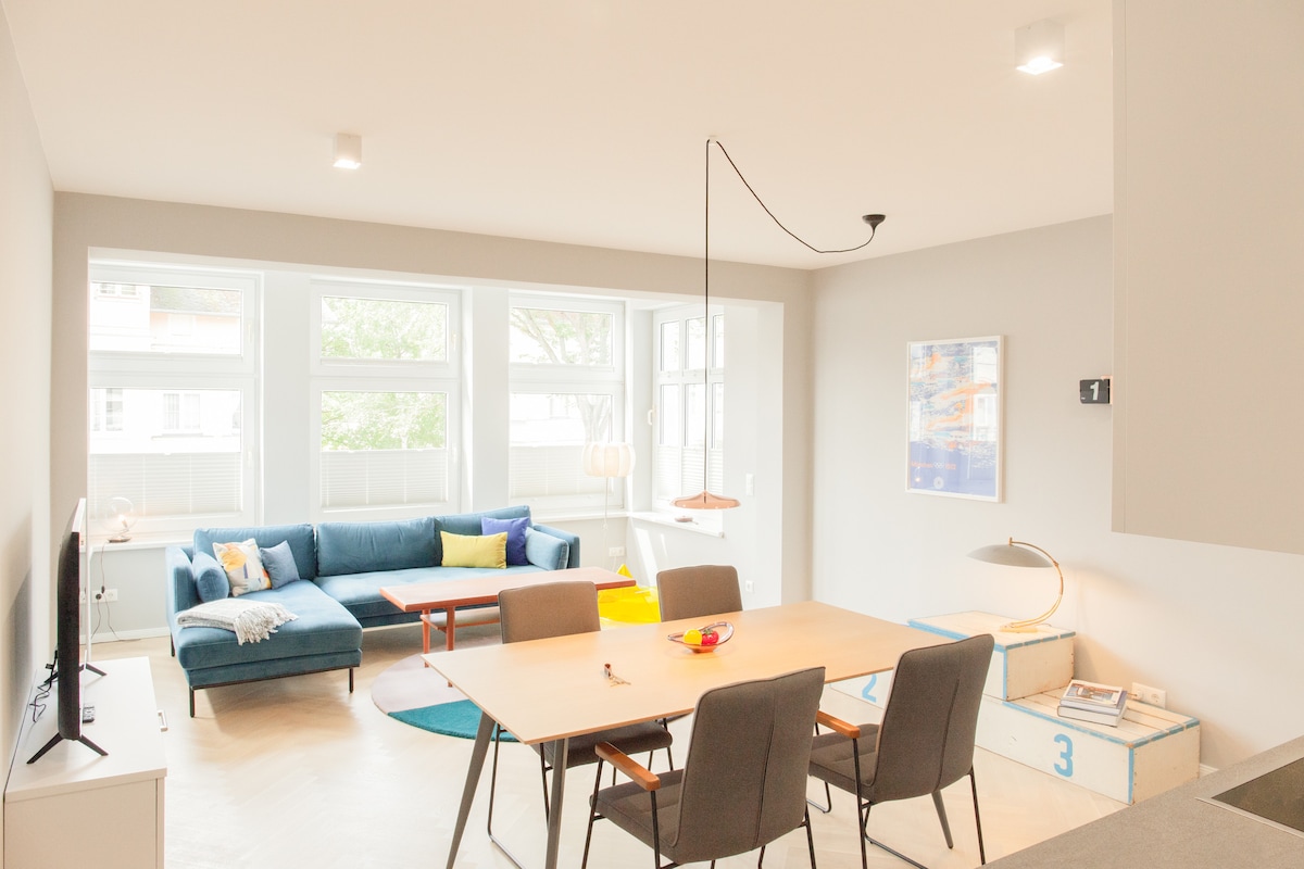 A bright living area features a sectional sofa in blue with accent pillows, a light wood dining table surrounded by gray chairs, and a light fixture above. Large windows provide natural light, with greenery visible outside, creating a spacious and welcoming environment.