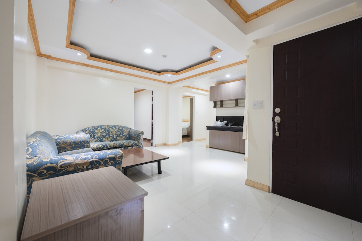 An inviting living area is shown with a decorative couch featuring blue and gold patterns. A wooden coffee table sits at the center, and the kitchen area is visible in the background, equipped with modern appliances. Natural light is provided by recessed ceiling lights.