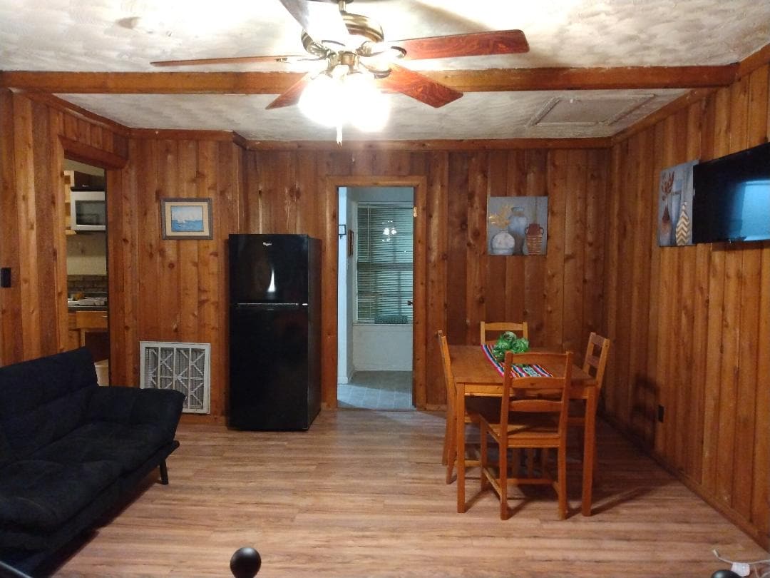 A cozy living area features wooden paneling and a ceiling fan. A dark couch is positioned near a small dining table with chairs. In the background, a doorway leads to a kitchenette with a refrigerator and an exit to a bathroom.
