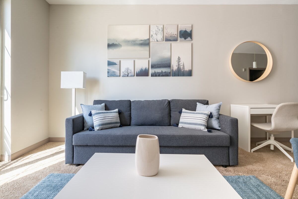 A modern living area features a gray sofa adorned with striped cushions. A round mirror is mounted on the wall above a white coffee table with a decorative vase. An abstract artwork hangs on the wall, while natural light fills the space from large windows.