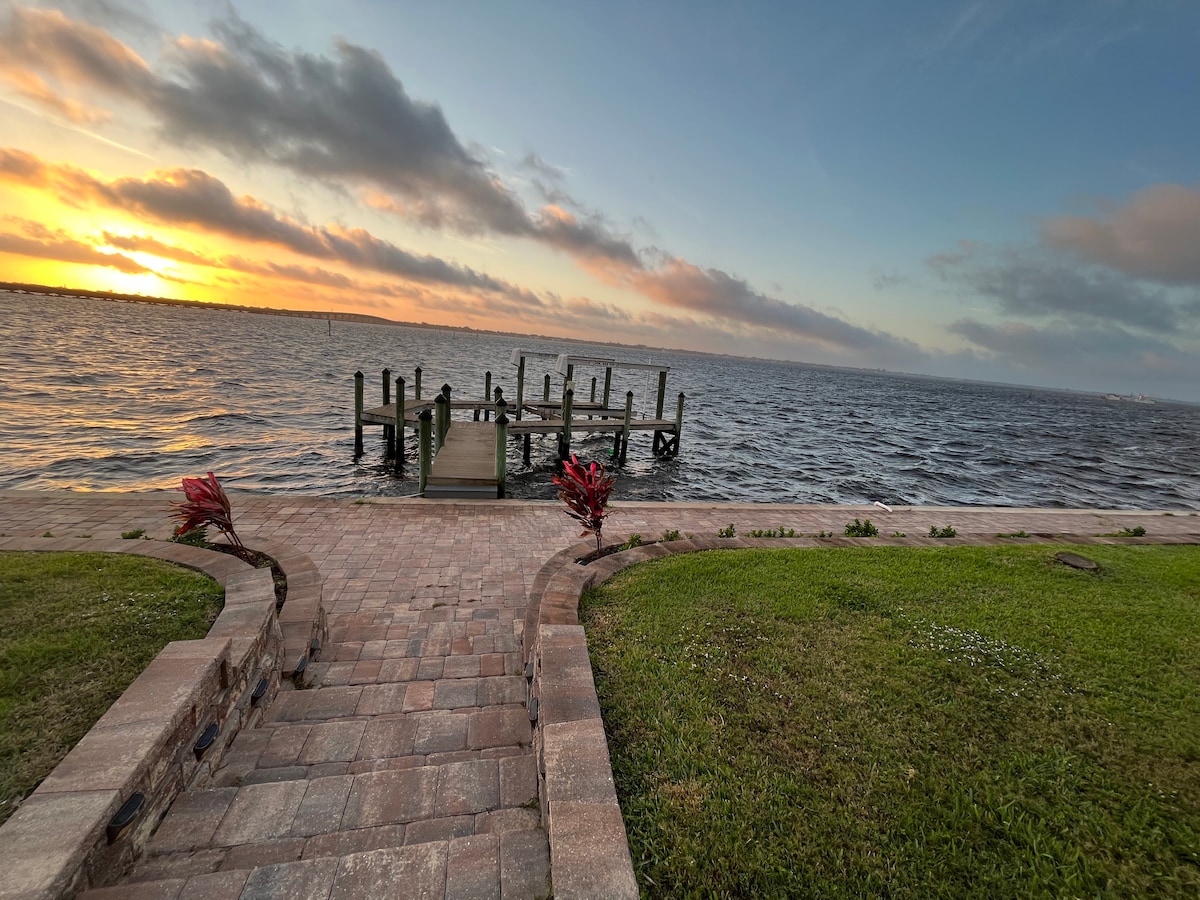A serene waterfront pathway is lined with lush green grass and vibrant plants, leading to a private dock. The calm waters reflect the soft colors of the sunset, while clouds drift lazily across the sky, creating a peaceful ambiance.