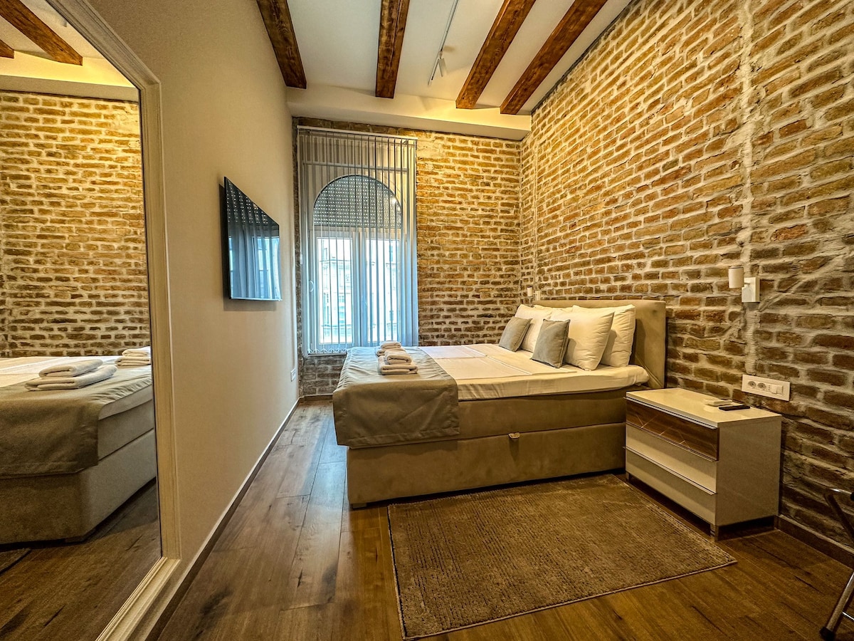 A cozy bedroom features exposed brick walls and wooden beams overhead. A spacious bed is adorned with neatly arranged linens and towels. Natural light filters through the large window, enhancing the warm ambiance. A large mirror reflects the inviting space, while a bedside table offers minimalist storage.