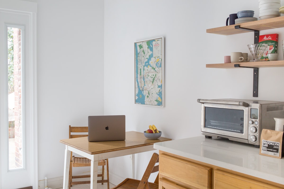A bright kitchenette features a wooden dining table paired with a chair, complemented by a laptop on the surface. Open shelves display a map of the area, dishes, and kitchenware, while a countertop oven is positioned beside a full-size fridge. Natural light enters through a nearby window.