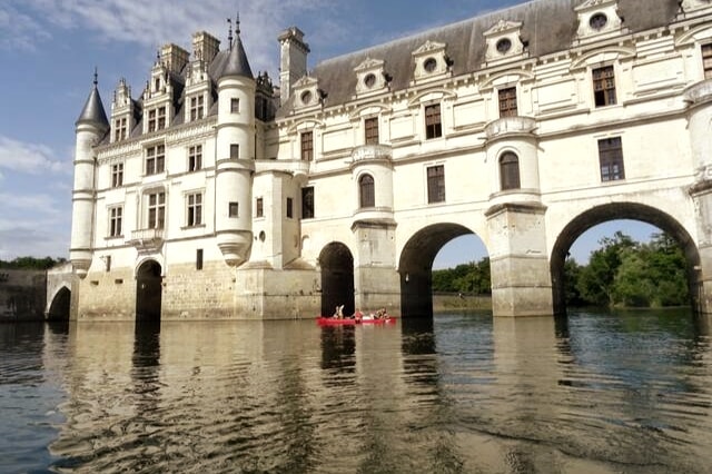 A historic chateau stands majestically by the water, its architectural details reflecting in the calm surface. Two kayakers paddle leisurely beneath the grand arches, surrounded by lush greenery in the background. The sky is clear, enhancing the tranquil scene.