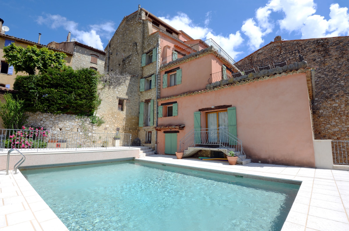 A shared swimming pool is surrounded by a tiled deck, offering a refreshing space for relaxation. The building's exterior features stone and pastel-colored walls, with several large windows allowing natural light. Lush greenery and blooming flowers enhance the setting.