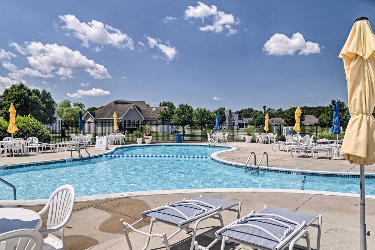 An outdoor pool is surrounded by lounge chairs and umbrellas, providing ample seating for relaxation. The clear water reflects the sky, while well-kept green spaces and community buildings are visible in the background. The area is open and inviting, ideal for leisure.