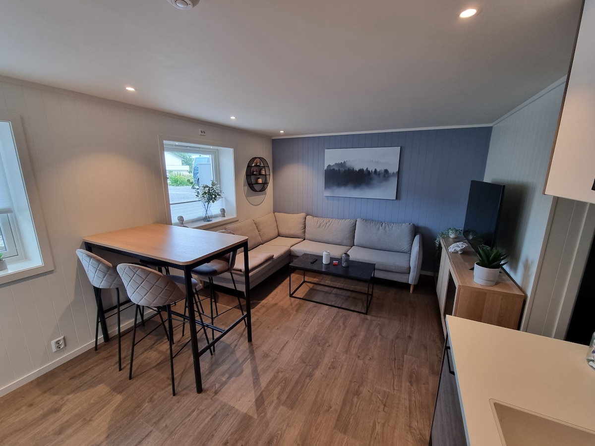 A modern living area showcases a spacious, gray sectional sofa arranged around a black coffee table. A dining table with four chairs is positioned adjacent. Soft lighting enhances the light-colored walls, while large windows allow natural light to fill the space.