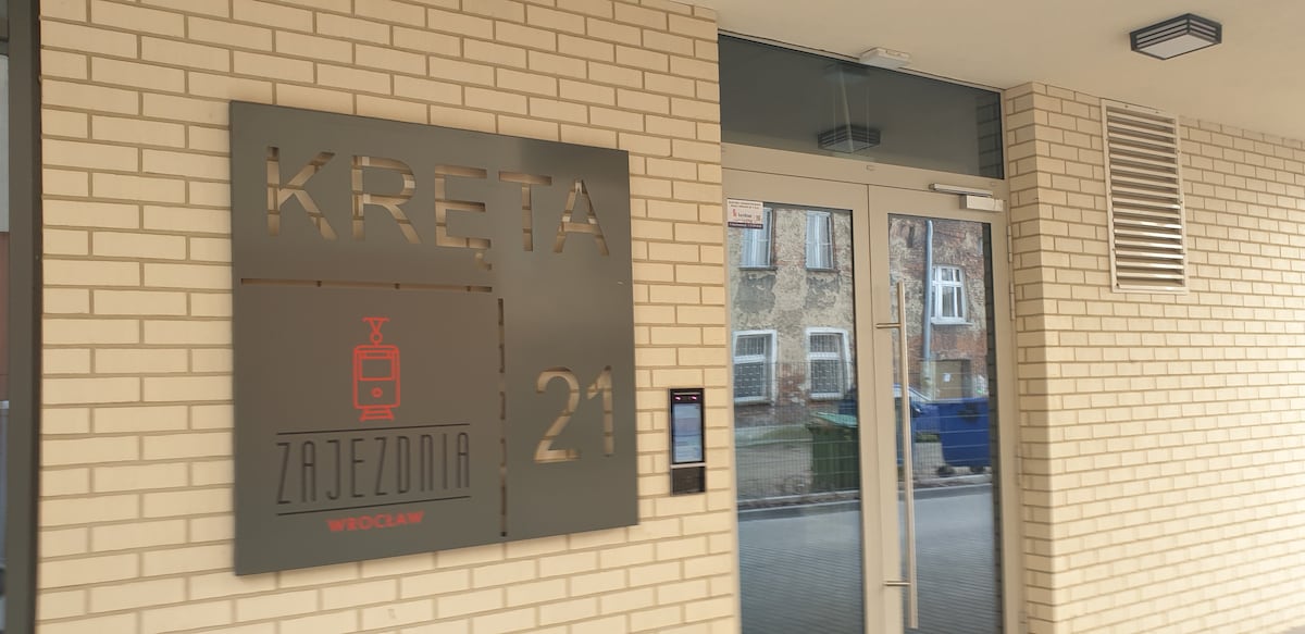 A modern entrance sign is displayed on a cream-colored brick wall, featuring the name 'KRETA 21' in stylized lettering. Glass double doors provide access to the building, with intercoms and a directory visible alongside.