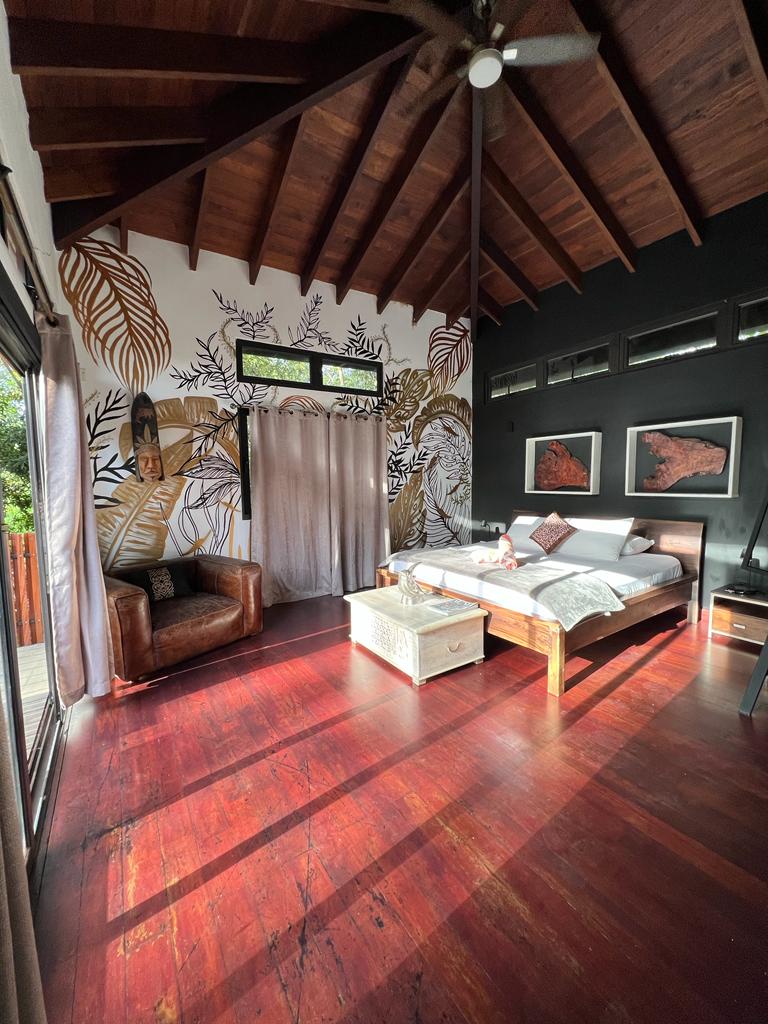 A spacious room is adorned with a high wooden ceiling and a striking mural. A large bed is positioned thoughtfully in the center, complemented by a cozy armchair. Natural light filters in through expansive windows, casting warm shadows across the polished wooden floor.