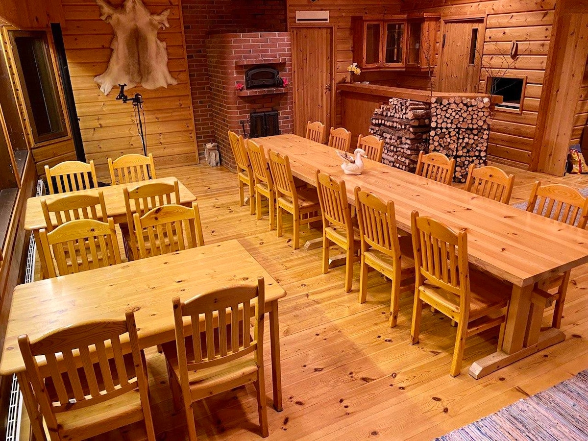 A spacious common room features warm wooden walls and flooring. Two large wooden tables, accompanied by matching chairs, occupy the space. A cozy fireplace is positioned against the back wall, with neatly stacked logs nearby. Large windows allow for natural light to brighten the area.