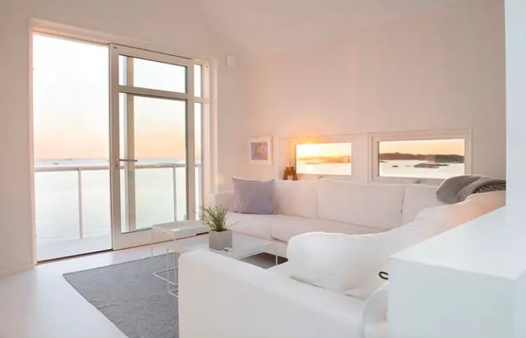 A modern sitting area features a light-colored sectional sofa positioned near large glass doors. Natural light floods the space, highlighting the views of the ocean and the sunset. A minimalist coffee table sits on a soft rug, completing the serene atmosphere.