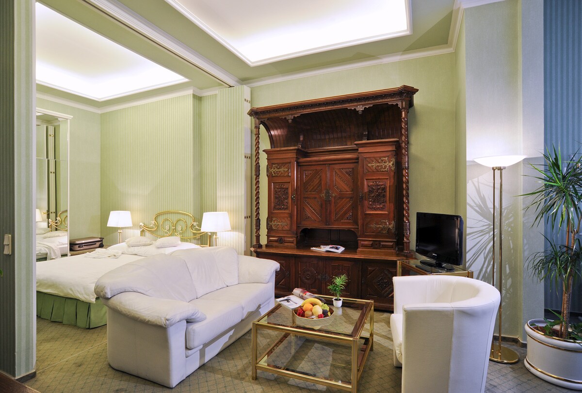 A spacious room features elegant green walls and soft lighting. A large bed is positioned near an intricately carved wooden cabinet. A white armchair sits beside a small coffee table adorned with fresh fruit, while a tall plant adds a touch of nature to the ambiance.