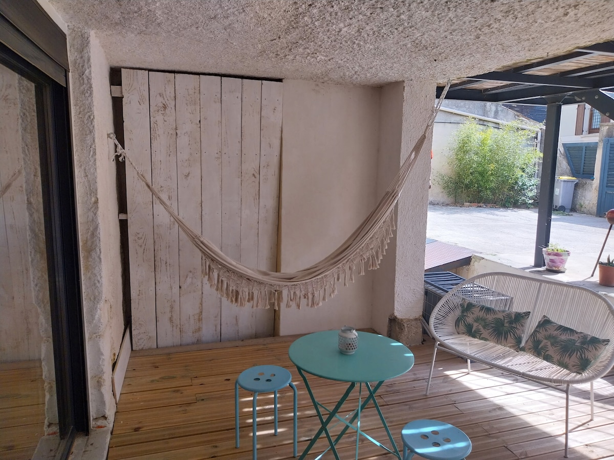 A relaxing terrace space features a light-colored hammock suspended on one side, inviting leisure. A small turquoise table accompanies two white chairs with floral cushions, while a wooden floor adds warmth. Natural light fills the area, complemented by greenery visible in the background.