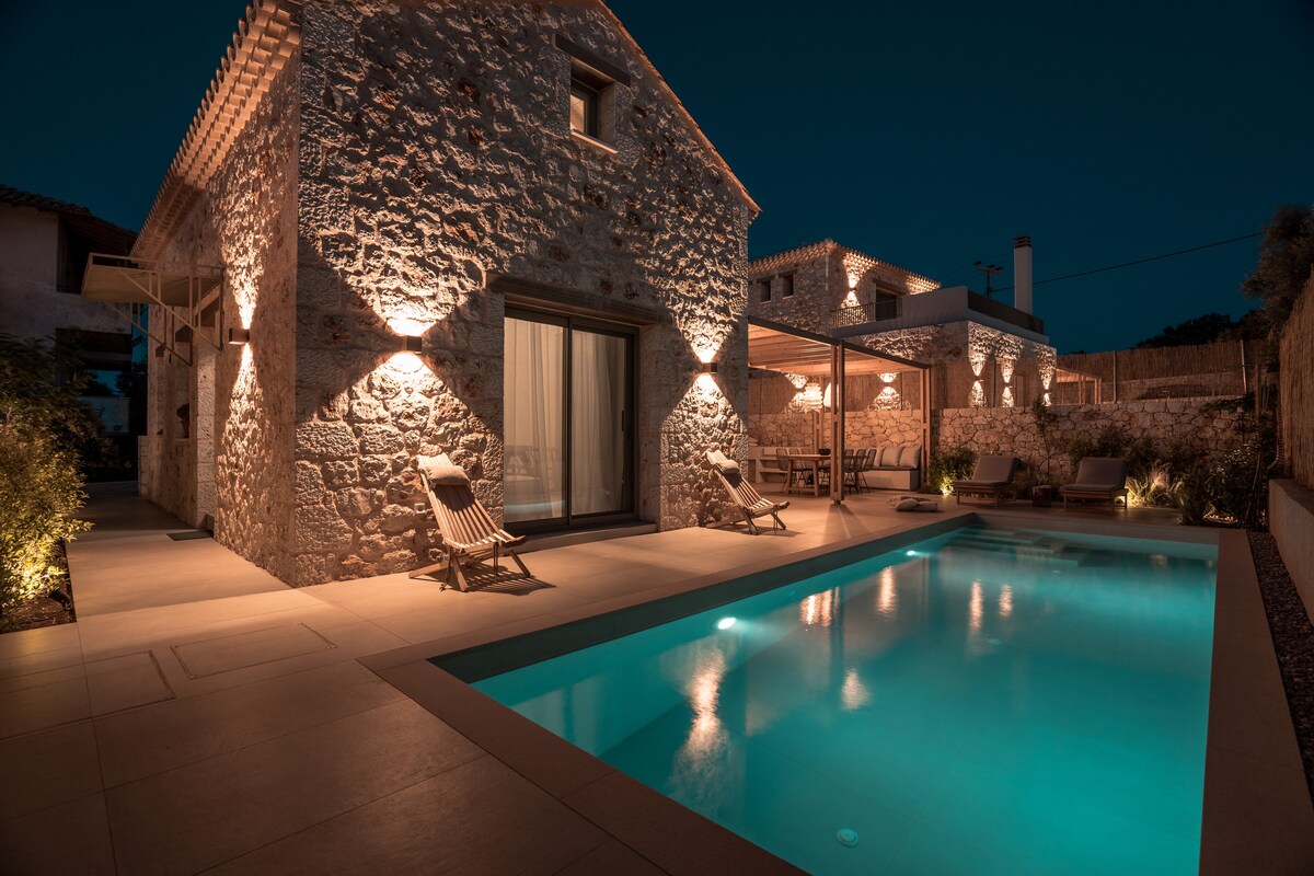 The exterior of the villa is softly illuminated at night, highlighting the traditional stone walls. A private swimming pool reflects the glow of outdoor lighting, while a shaded patio area offers comfortable seating options near the pool.
