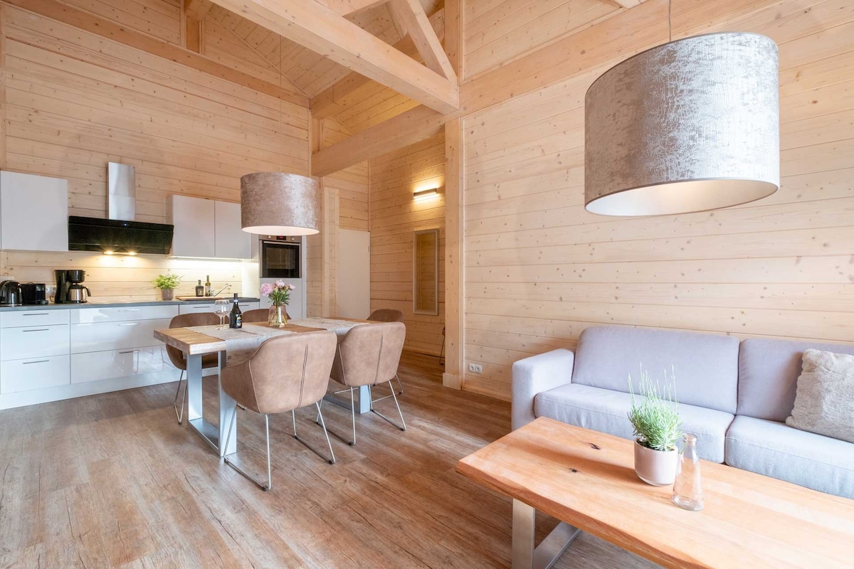 An open living space features light wood paneling and high ceilings. A stylish dining table is paired with comfortable chairs, while a plush sofa adds softness. Modern kitchen appliances and a decorative table centerpiece complete the inviting atmosphere.