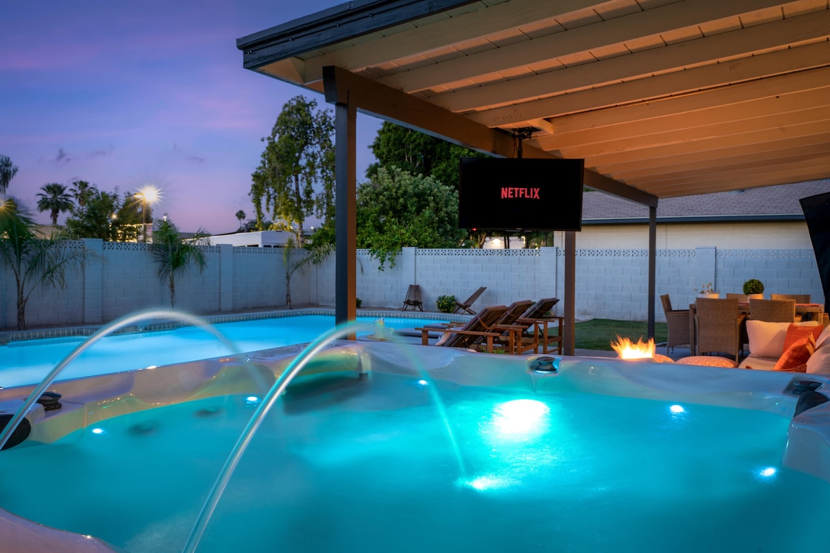 The hot tub is illuminated in calming turquoise light, featuring fountain jets that add a soothing touch. A mounted screen displays the Netflix logo, while comfortable loungers are positioned nearby under the shaded patio, offering a relaxing atmosphere for outdoor enjoyment.