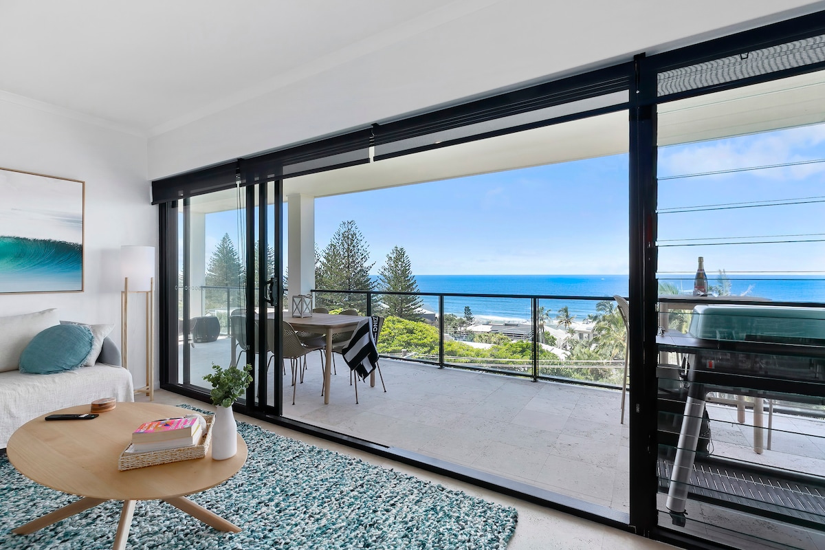 A spacious living area features large sliding glass doors that open to a balcony, providing panoramic views of the ocean. A round coffee table sits on a textured rug, accompanied by a cozy sofa. A dining table is set for meals with chairs arranged for gatherings.