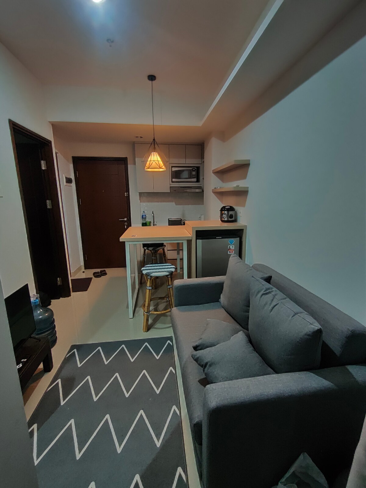 A compact living space presents a cozy seating area with a gray sofa and a patterned rug. A small dining table is positioned next to a kitchenette, featuring a microwave and refrigerator. Soft lighting is provided by an overhead pendant light.