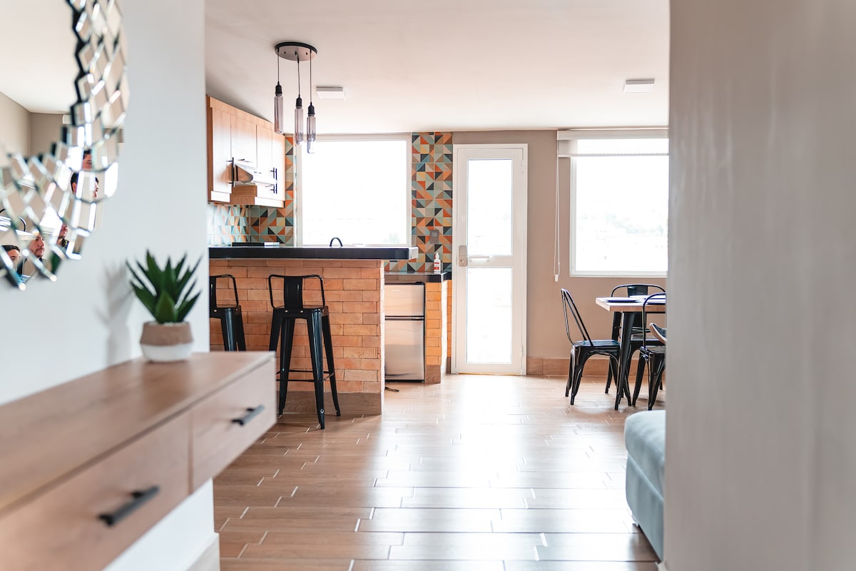 An inviting living space features a modern kitchen with wooden cabinets and a colorful tiled backsplash. High-top black stools are positioned at a counter, while a dining area includes black chairs around a table. Natural light floods in from large windows, enhancing the open layout.
