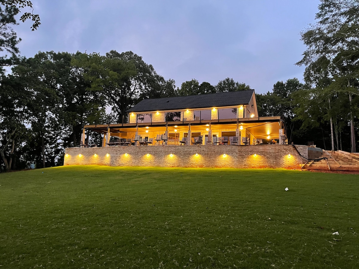 A spacious house is illuminated by outdoor lighting, showcasing a large wrap-around deck. The home is set against a backdrop of trees, with a well-manicured lawn leading up to it. Soft evening light creates a welcoming atmosphere around the property.
